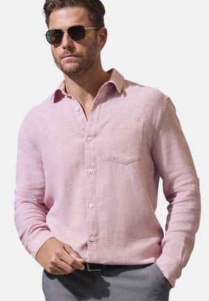 Man wearing black sunglasses and light pink long-sleeve button-up shirt tucked into gray pants, standing with one hand in pocket.