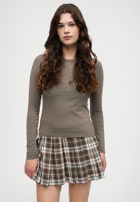 Μακρυμάνικο, εφαρμοστό ελαιόχρωμο μπλουζάκι συνδυασμένο με μια πλισέ φούστα σε καρό σχέδιο με καφέ, λευκό και μαύρο χρώμα.