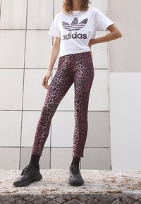 Person wearing white Adidas cropped T-shirt, purple leopard print leggings, black socks, and black sneakers, standing against tiled wall.