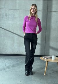 Henkilöllä päällään violetti pitkähihainen paita, mustat farkut ja mustat saappaat. Taustalla betoniseinä ja vaaleasta puusta valmistettu pöytä, jolla lehti ja kuppi.