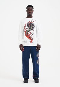Ein Modell posiert in einem weißen Langarmshirt mit buntem Drachendesign und blauen Jeans mit Stickereien, vor einem einfarbigen Hintergrund.