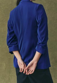 Blauwe blazer met gestructureerde schouders, lange mouwen opgerold bij de manchetten, en een gladde stof, gecombineerd met donkere broeken. Achteraanzicht.