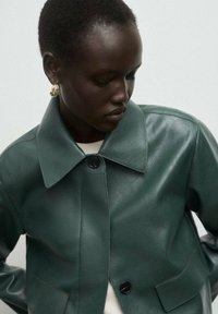 Chaqueta de cuero verde con frente abotonado, amplio cuello y dos bolsillos. Textura lisa con sutiles detalles de costura.