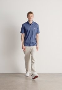 Modrý polo golier s krátkym rukávom s golierom, na ktorom je malý logo. Kombinovaný s krémovými nohavicami a bielymi teniskami na neutrálnom pozadí.