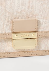 Beige käekott, millel on tekstureeritud lillemuster, kullatooni metallne klapp ja õmbluse detailid siledal alusel.