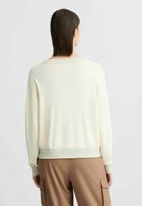 Maglione chiaro in crema con una vestibilità rilassata, polsini e orlo a coste. Presenta un ampio scollo e una texture morbida, abbinato a pantaloni cargo marroni.