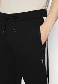 Svarta joggers med elastisk midja, dragsko, dragkedjefickor på sidan och grå sidskaror. Har ett broderat logotyp på vänster ben.