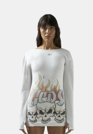 Žena oblečená v bílém tričku s dlouhým rukávem, na přední straně s grafikou tří lebek a plamenů a písmeny "GCDS".