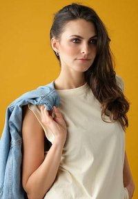 Top senza maniche di un beige chiaro con scollatura tonda. Giacca di jeans appoggiata sulla spalla. Sfondo giallo vibrante. Lunghi capelli castani e mossi.