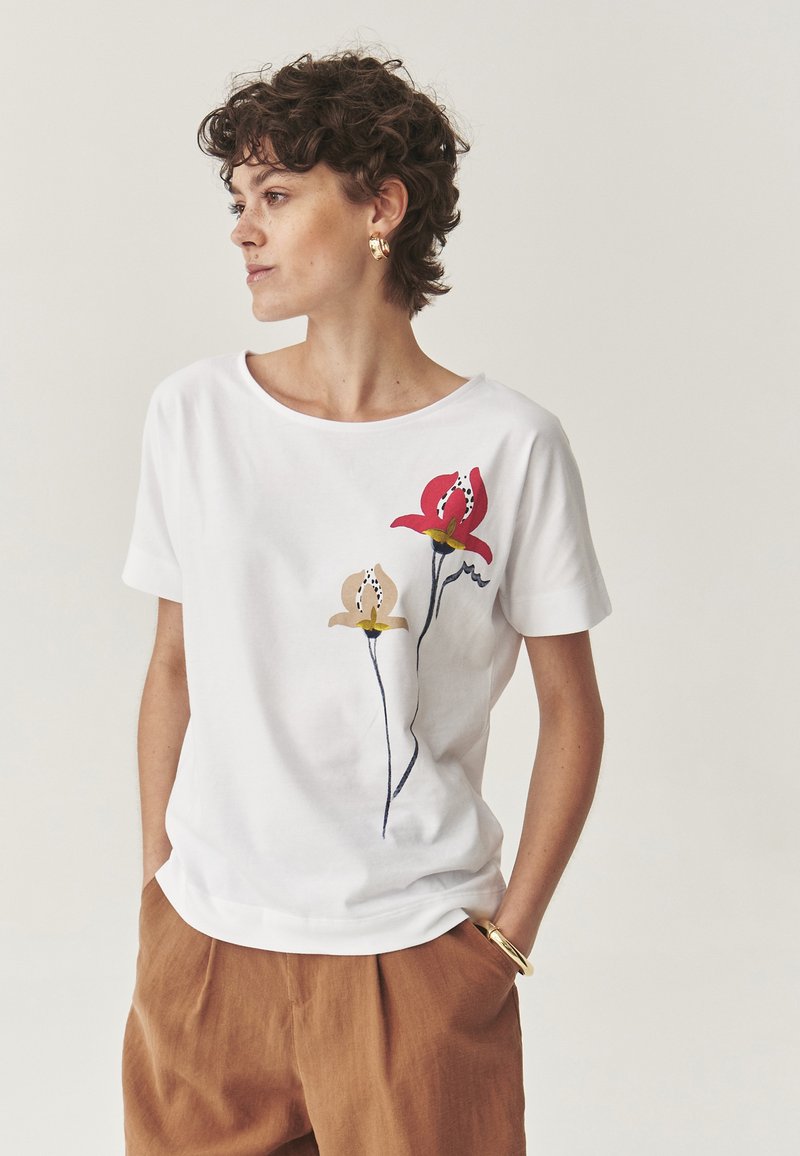 Balta medvilninė T-shirt marškinėliai su gėlių raštu, kuriame matoma raudona gėlė ir smėlio spalvos gėlė, abi ant ilgo stiebo. Trumpi rankovės ir plati kaklo iškirptė.