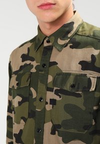 Camo-shirt met groene, beige en zwarte patronen; button-down kraag; twee borstzakken; zwarte knopen; textiel met textuur.