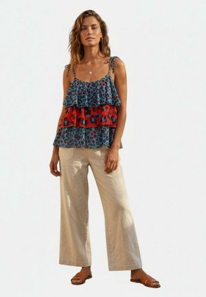 Donna che indossa un top smanicato multistrato blu e rosso con stampa leopardata, pantaloni beige a gamba larga, sandali marroni e una collana d'oro.