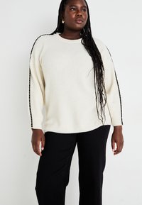 Weißer gestrickter Pullover mit seitlichen schwarzen Akzenten, lockerer Passform, Rundhalsausschnitt und langen Ärmeln, kombiniert mit schwarzen weiten Hosen.