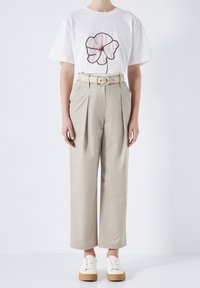 Personne portant un t-shirt blanc avec un imprimé floral, un pantalon beige taille haute plissé avec une ceinture, et des baskets blanches avec des semelles en caoutchouc.