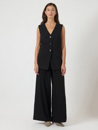 Γυναίκα ντυμένη με αμάνικο μαύρο γιλέκο με κουμπιά και φαρδύ μαύρο παντελόνι, φορώντας γκρι γόβες με μυτερή μύτη, στάσμένη μπροστά σε άσπρο φόντο.