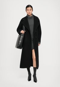 Femme portant un long manteau noir sur un pull col roulé gris et une jupe, portant un grand sac fourre-tout noir, se tenant devant un fond blanc.