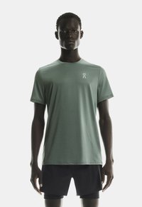 Bărbat îmbrăcat cu un tricou sport cu mânecă scurt�ă de culoare verde deschis și pantaloni scurți sport negri, stând în fața unui fundal alb simplu.