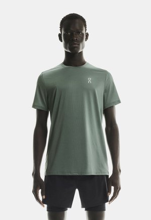 Hombre vistiendo una camiseta atlética de manga corta verde claro y pantalones cortos atléticos negros, de pie frente a un fondo blanco simple.