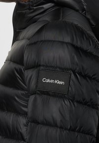 Sort pufferjakke med quiltet design, der har et stofmærke med "Calvin Klein" tekst på venstre skulder. Glat, skinnende tekstur.