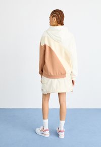 Sweatshirt à capuche en crème et terre cuite, en tissu texturé avec des blocs de couleur diagonaux. Assorti à une jupe beige clair et des baskets blanches.