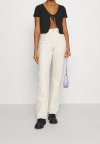 Cardigan noir côtelé court avec des bords festonnés, associé à un pantalon large taille haute crème. Le modèle tient un petit sac à main lavande.