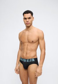 Άνδρας σε φόρμα στέκεται προς τα εμπρός φορώντας μαύρα εσώρουχα Calvin Klein με φωτεινό μπλε λογότυπο στη ζώνη της μέσης, σε απλό ανοιχτό φόντο.