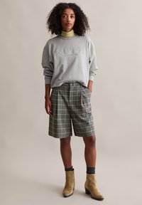 Frau mit lockigem Haar, die einen grauen Sweatshirt, einen gelben Rollkragenpullover, graue karierte Shorts, dunkle Socken und beigefarbene Wildleder-Stiefeletten trägt und vor einem einfarbigen Hintergrund steht.