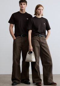 Μαύρα T-shirt και καφέ παντελόνια φαρδιάς γραμμής, με μικρό λογότυπο, συνδυασμένα με κρεμ τσάντα χειρός και μαύρα-βυσσινί παπούτσια.