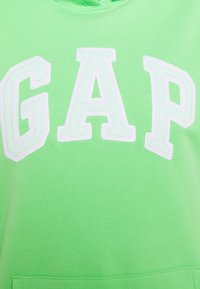 Zelená mikina s kapucí vyrobená z měkké tkaniny má výrazné logo "GAP" v bílé barvě s texturovaným vyšitým povrchem. Obsahuje přední kapsu.