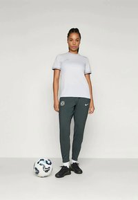 Svijetlosivi sportski majica kratkih rukava sa suptilnim logotipom, uparena s tamnozelenim suženim trenirka hlačama koje također imaju logotip, i crnim sportskim cipelama.