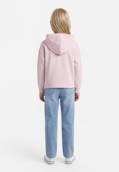 Otrok z dolgimi blond lasmi, oblečen v svetlo roza kapucar, modre kavbojke in bele superge, stoji z obrazom stran od gledalca na belem ozadju.