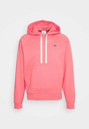 Sweat à capuche rose corail avec poche avant, cordons blancs, poignets côtelés et petit logo crocodile Lacoste vert sur la poitrine.