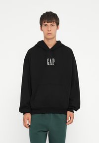 Sudadera con capucha negra que presenta un bolsillo frontal y la marca "GAP" en blanco. Fabricada con una tela suave, tiene un corte relajado y puños acanalados.