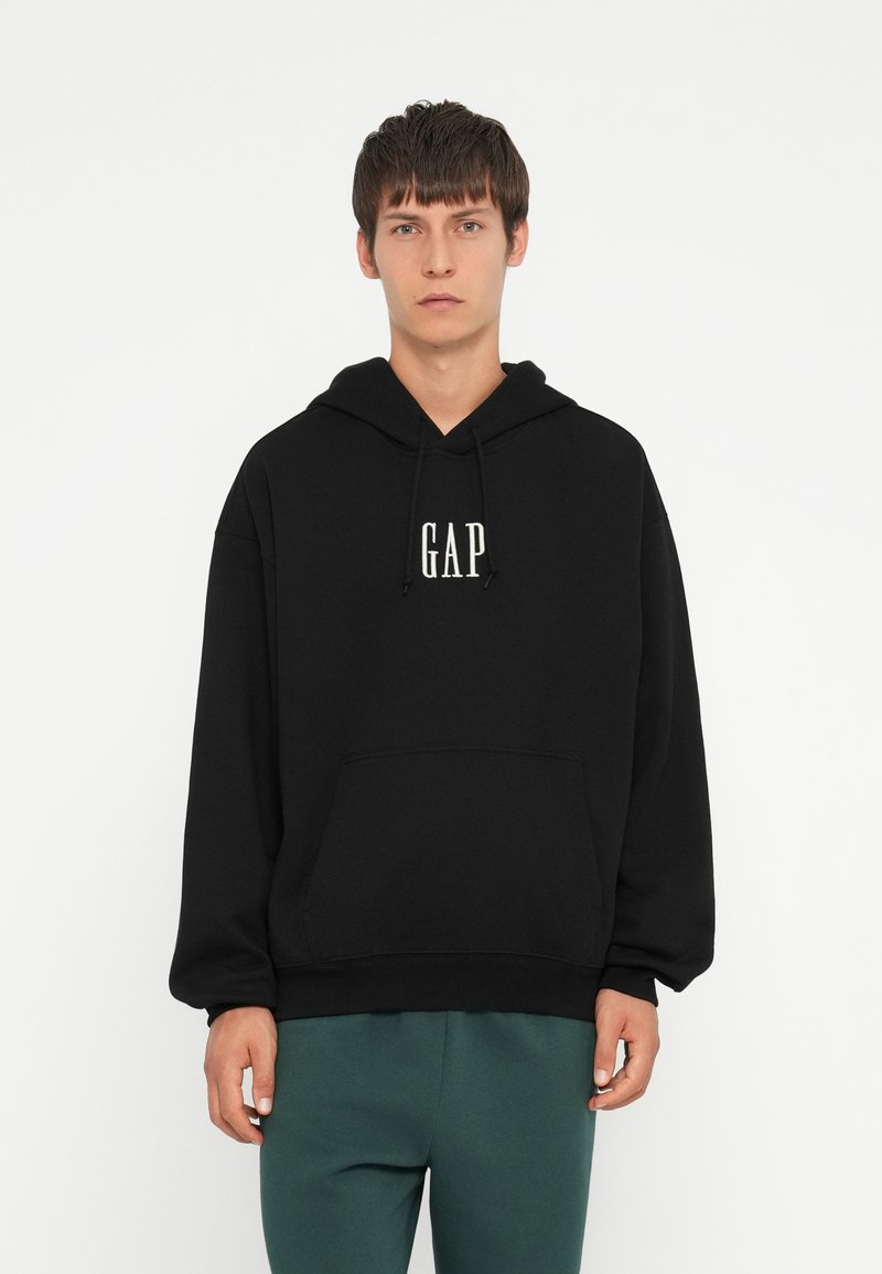 Sudadera con capucha negra que presenta un bolsillo frontal y la marca "GAP" en blanco. Fabricada con una tela suave, tiene un corte relajado y puños acanalados.