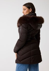 Cappotto imbottito marrone con cappuccio bordato di pelliccia, caratterizzato da sezioni trapuntate diagonali e un design a vita adatta. Indossato sopra jeans azzurri chiari.