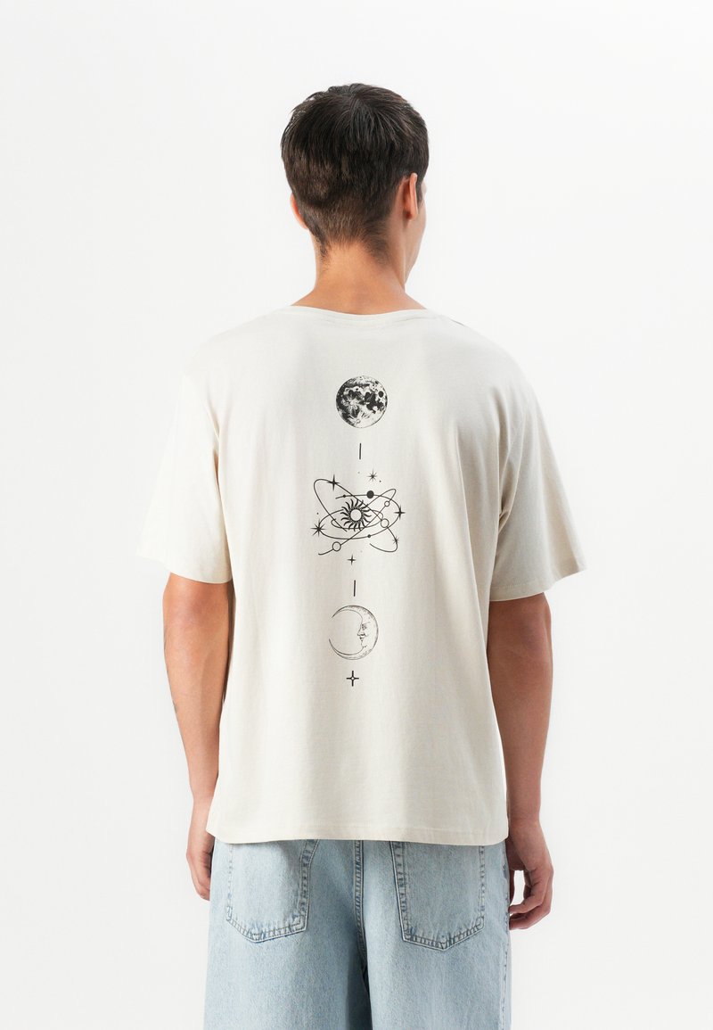 Man wearing off-white t-shirt with black celestial design of moon, sun, and stars on back, paired with light blue denim shorts.
