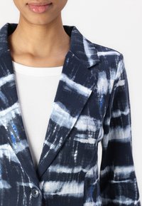 Veste à motifs bleu marine et blanc avec un tissu texturé, des revers crantés et une fermeture à un bouton. Portée sur un haut blanc côtelé.