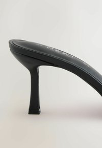 Mule sandale en cuir noir à talon haut fin avec un talon aiguille et une semelle intérieure légèrement rembourrée sur un fond neutre.
