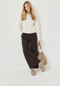 Felpa cappuccio beige chiaro, pantaloni ampi marroni abbinati, sneakers beige con suola rigata, e una borsa marrone morbida.