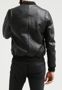 Mand iført sort læderjakke og sorte jeans, set fra ryggen mod en ensfarvet lys baggrund.