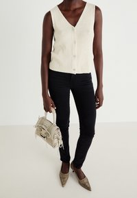 Krämfärgad stickad väst med V-ringning och knäppning, ihop med smala svarta jeans och metallspetsade klackar, hållande en liten beige väska.