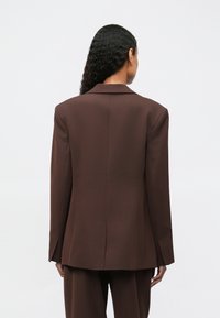 Woman with long curly hair facing away, wearing a loose, dark brown tailored blazer and matching trousers against a plain background.