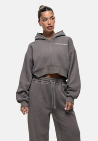 Hoodie crop in tessuto grigio con zip frontale, cappuccio e maniche larghe. Presenta il testo "GOODFORNOTHING" sul petto, abbinato a pantaloni coordinati.