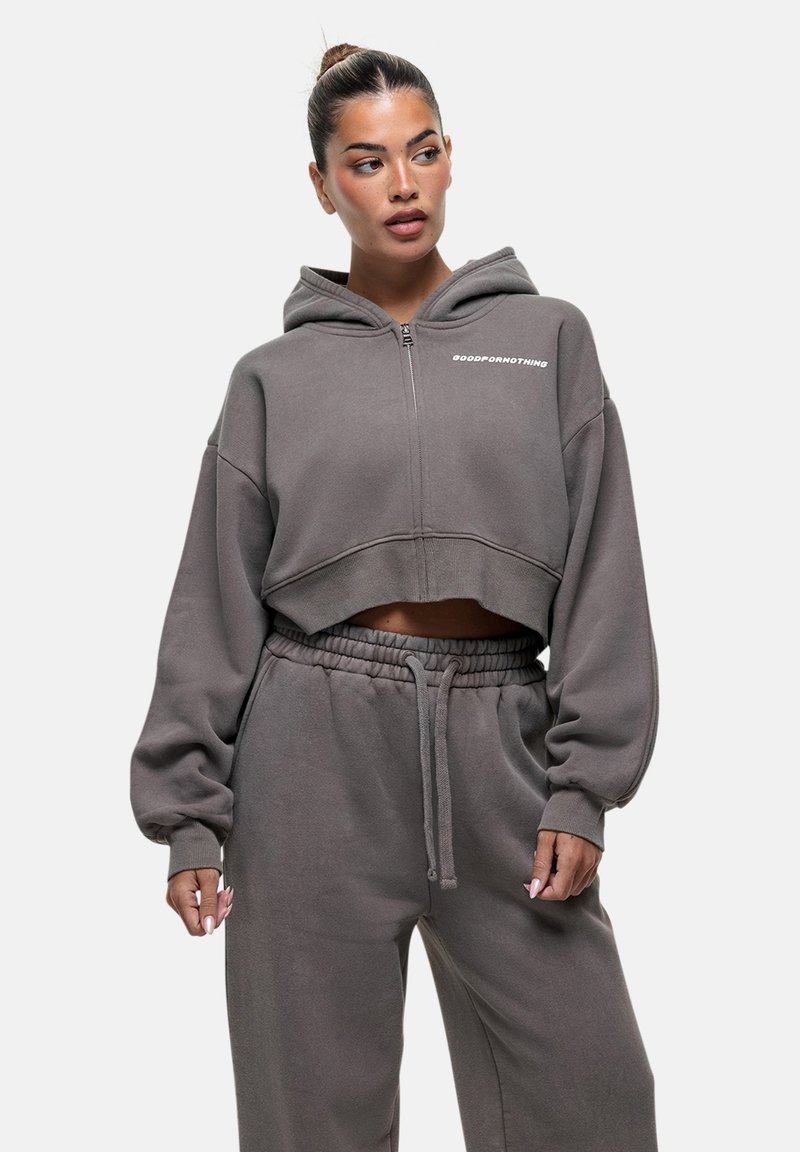 Hoodie crop in tessuto grigio con zip frontale, cappuccio e maniche larghe. Presenta il testo "GOODFORNOTHING" sul petto, abbinato a pantaloni coordinati.