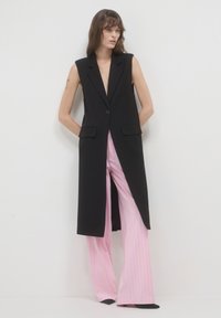 Schwarze ärmellose Blazer mit tiefem Ausschnitt, mit einem Knopfverschluss, zwei frontalen Taschen und taillierter Passform, kombiniert mit rosa gestreiften Hosen.