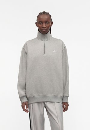 Dorosły noszący jasnoszary sweter z suwakiem do ćwiartki i logo Adidas oraz jasnoszare spodnie na jednolitym białym tle.