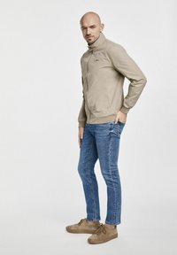 Beżowa bomberka z wysokim kołnierzem, zamek błyskawiczny i boczne kieszenie. Zestawiona z niebieskimi Skinny jeans i brązowymi zamszowymi trampkami.