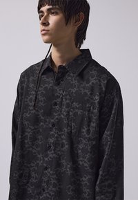 Camicia nera con bottoni, caratterizzata da un motivo scuro ripetuto, maniche lunghe, colletto e una tasca sul petto. Realizzata in un tessuto morbido e con texture.