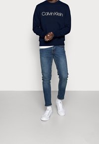Felpa navy con logo "Calvin Klein", abbinata a jeans blu e sneaker bianche. Design semplice, vestibilità casual. Texture morbide in tutto.
