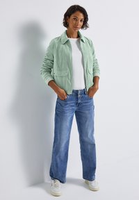 Grüne Cordjacke mit Kragen, zwei frontalen Taschen, kombiniert mit blauen Jeans und weißen Turnschuhen. Einfaches Design, strukturierter Stoff.